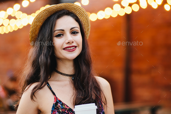 Pleased female with black hair, shining eyes and red lips, wearing straw hat and dress
