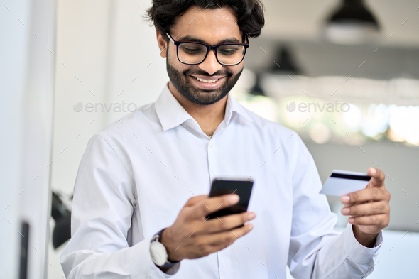Happy indian business man holding phone and credit card using mobile bank app.