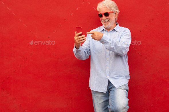 Senior handsome white-haired man standing over red background holding phone