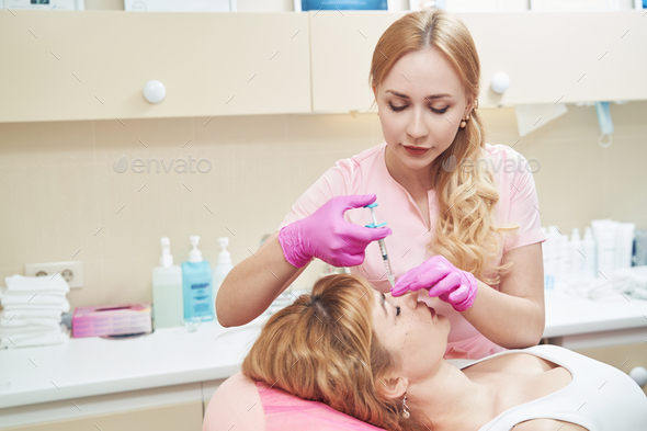Female beautician in uniform is doing procedure with filler in beauty centerm