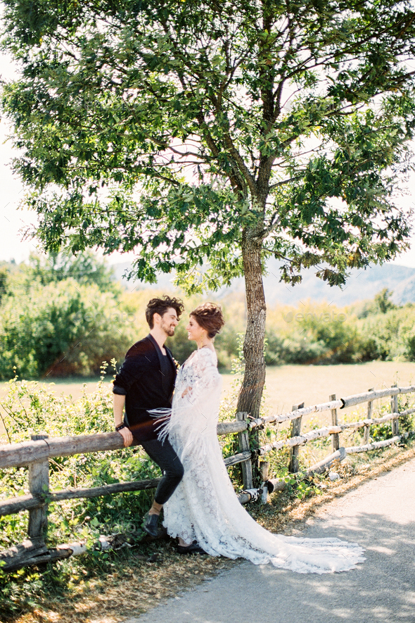 Bride stands near groom sitting on a wooden fence near a country road