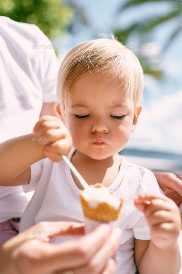 Little girl carefully takes ice cream with a spoon, sitting on mom lap