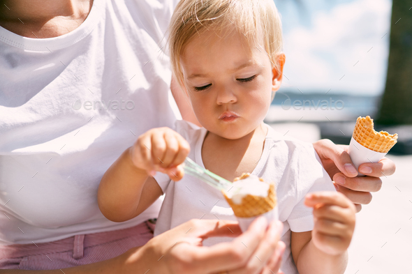 Little girl picks up ice cream with a spoon while sitting on mom lap