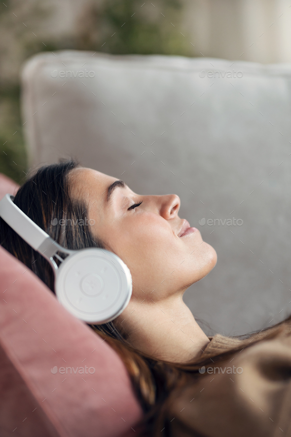 Beautiful woman listening to music with headphone while using smartphone lying on sofa at home. - Stock Photo - Images