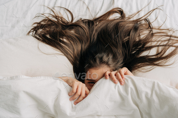 Young funny brunette woman is hiding under the covers while relaxing in bed.