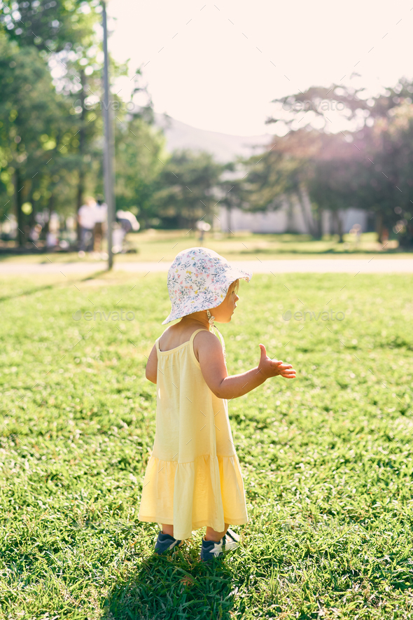 Little girl in a hat stands on the lawn with a raised hand. Side view