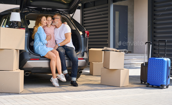 Daughter hugging smiling parents sitting in car with open trunk