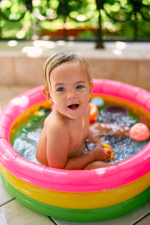 Cute wet baby with open mouth sits in inflatable mini pool, turning back