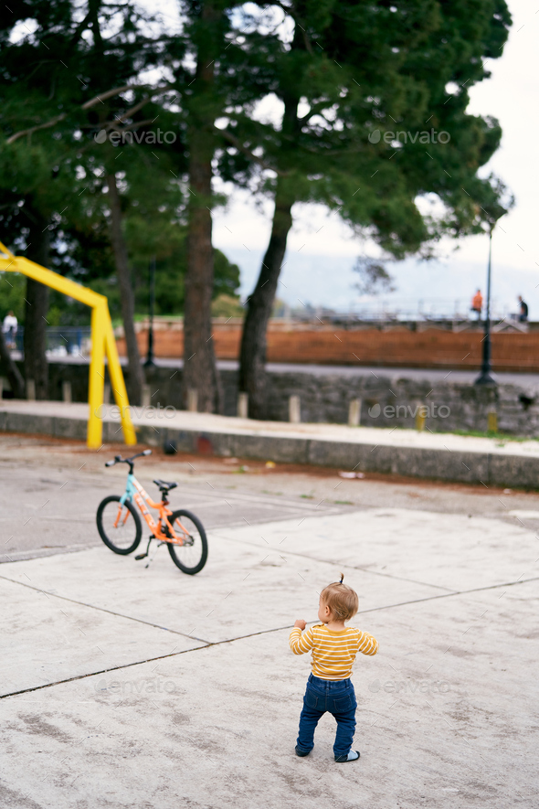 Little girl stands on a tiled playground in front of a bicycle in a park