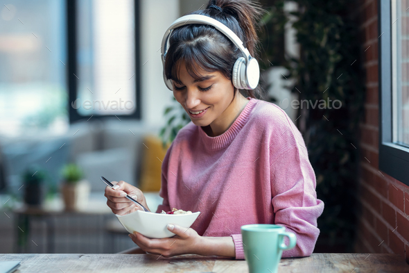 Beautiful young woman listening music with headphones while having healthy breakfast at home.