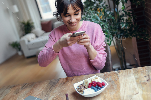 Beautiful young woman using her mobile phone while having healthy breakfast at home. - Stock Photo - Images