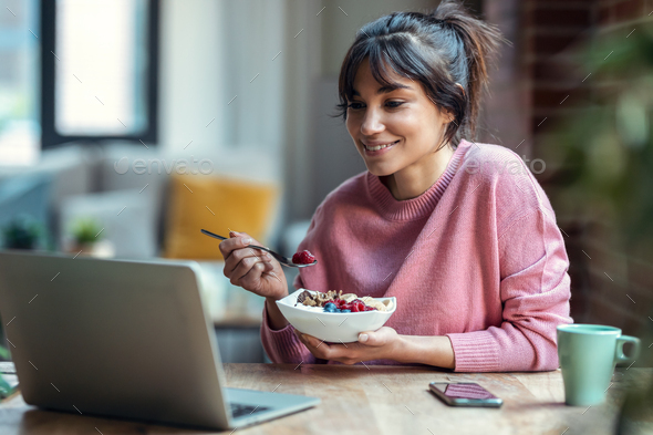 Beautiful young woman working with laptop while eating yogurt with fruits bowl at home. - Stock Photo - Images