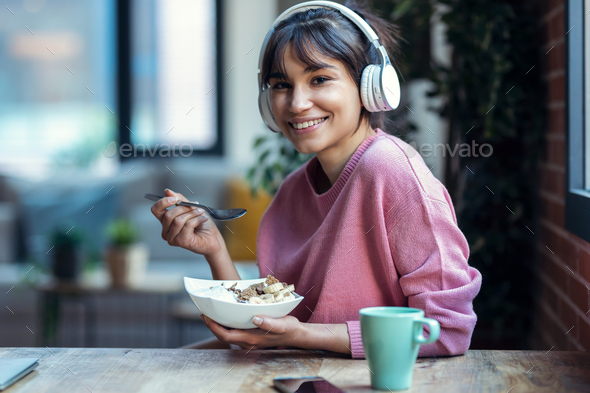 Beautiful young woman listening music with headphones while having healthy breakfast at home. - Stock Photo - Images