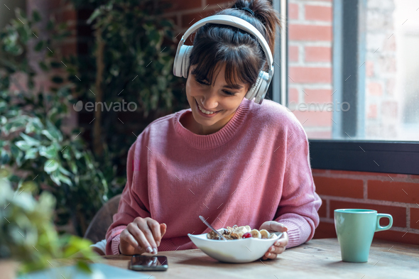Beautiful young woman listening music with headphones while having healthy breakfast at home. - Stock Photo - Images