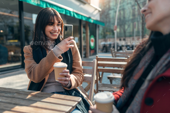 Two attractive friends enjoying coffee while taking a photo with smartphone sitting on a terrace - Stock Photo - Images