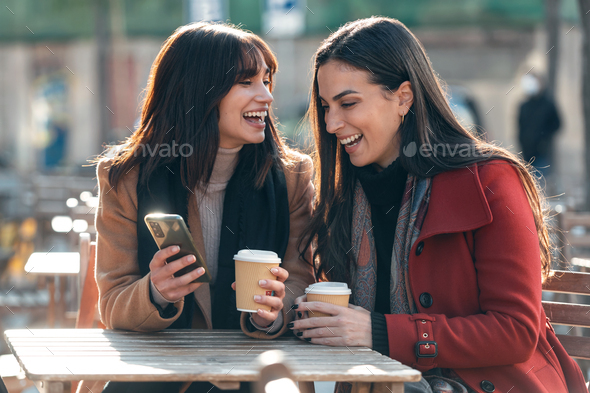 Two attractive friends enjoying coffee while looking smartphone and talking sitting on a terrace - Stock Photo - Images
