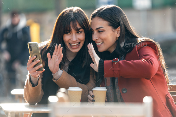 Two attractive friends enjoying coffee while doing a videocall with smartphone sitting on  terrace - Stock Photo - Images