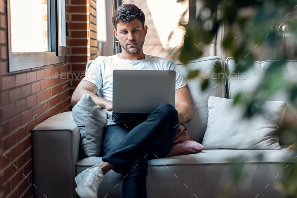 Smart man working with her laptop while sitting on a couch at home - Stock Photo - Images