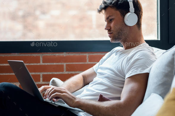 Smart man working with her laptop while listening music with headphones sitting on a couch at home - Stock Photo - Images