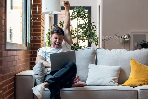 Successful young man using her laptop and celebrating something while sitting on sofa at home. - Stock Photo - Images