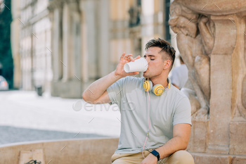 Young studen guy with headphones sits on street and drink cofffee in takeaway cup