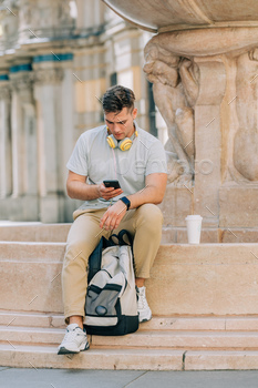 Young student guy with backpack sits on city street and using mobile phone.