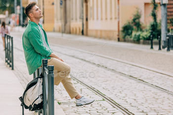 Stylish student guy in green shirt sits on railing on street