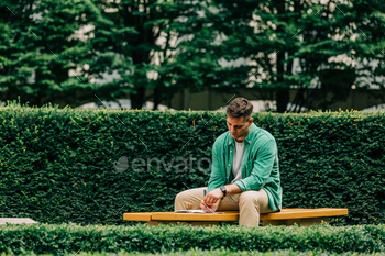 Stylish student guy in green shirt sits on bench in garden and write something