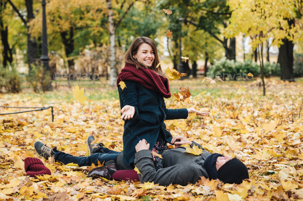 Positive woman wears knitted scarf, sits on boyfriend, throws yellow leaves in air