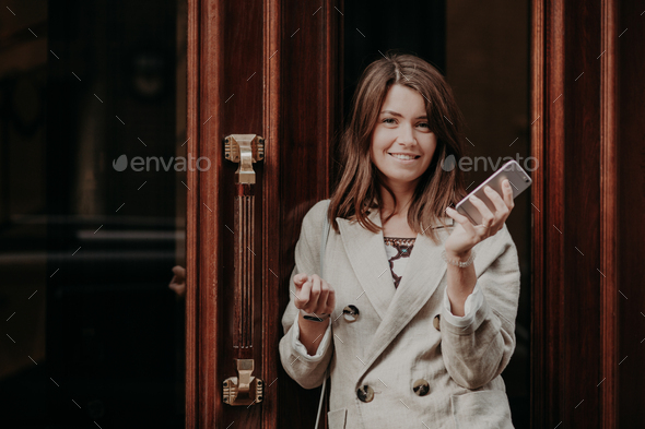 Elegant gorgeous lady in raincoat, holds mobile phone, waits for call, poses outdoor near doors