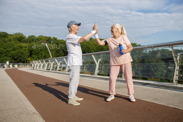 Joyful senior man and woman in sportswear give five training together on footbridge