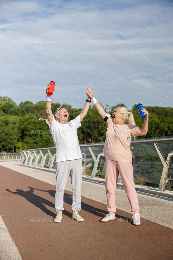 Joyful mature couple in tracksuits raises up bottles and gives five standing on city footbridge