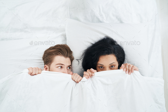 Young couple hiding their faces behind duvet