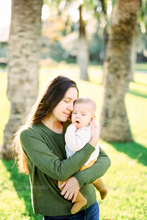 Mom hugs a sleepy baby in her arms, standing near palm trees