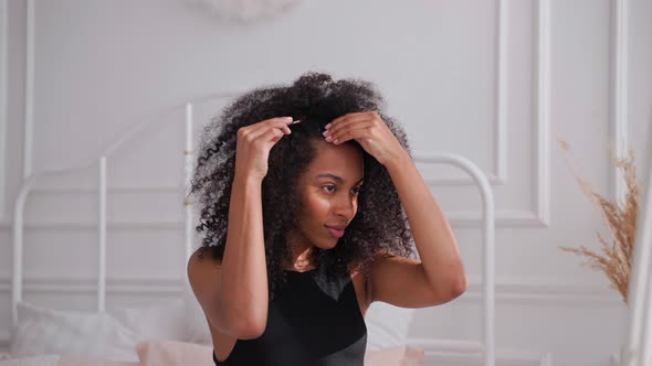 Black Woman Combing Her Afro Hair Sitting on a Bed and Looking in Mirror alt