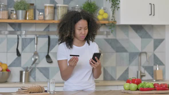 African Woman Making Online Payment on Smartphone in Kitchen alt