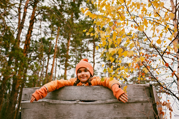 Happy girl in orange coat walks and plays in handmade wooden house in autumn park. Active weekend