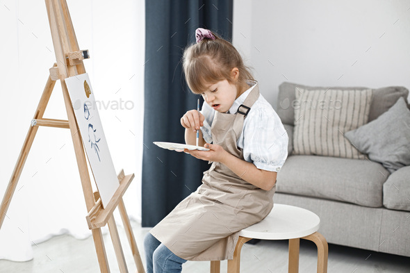 Girl with Down syndrome wearing beige apron and painting on an easel