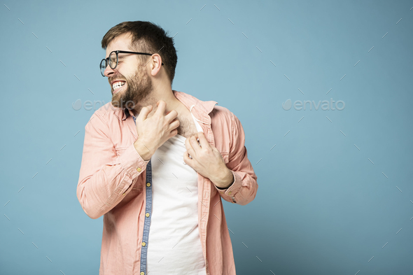 Tense man in glasses scratches itchy skin on neck with hand, painfully grins teeth and closes eyes