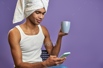 Young transgender man in towel on head with cup of tea and mobile in hands