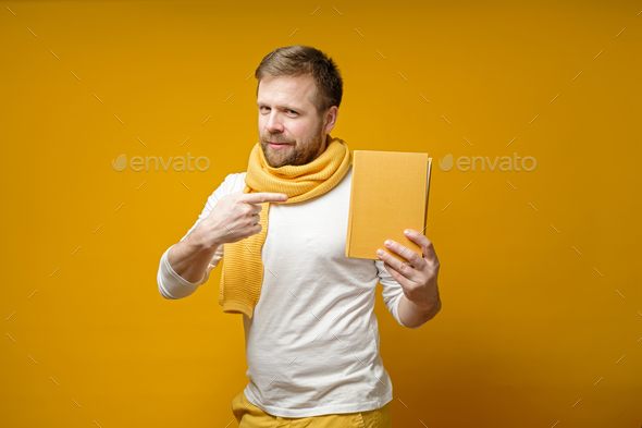 Cute man in a scarf, holds a yellow book in hands, points to it with index finger