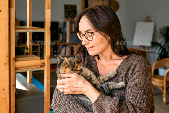 Middle-aged woman hugging cute tabby cat in indoor scene. Human-animal relationships. Funny home pet