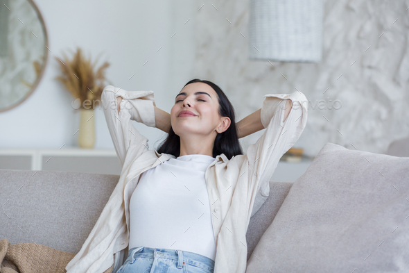 Happy young beautiful woman relaxing at home on sofa with eyes closed, arms behind head