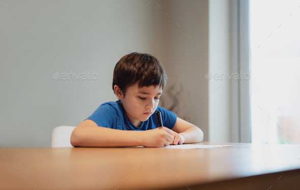 Kid siting on table doing homework,Child boy holding black pen writing on white paper,