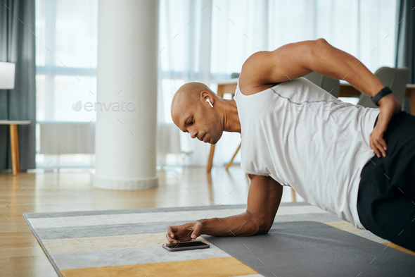 Black athletic man in side plank pose using smart phone while exercising at home.