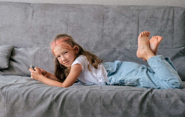 Little girl lying on couch and using smartphone. Concept of children\'s gadgets addiction.