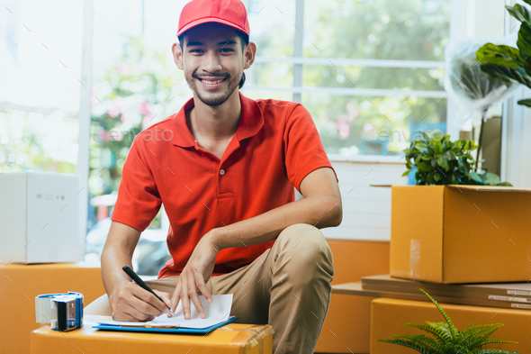 Professional Asian deliveryman in red uniform smile and check cargo box and parcel in home office