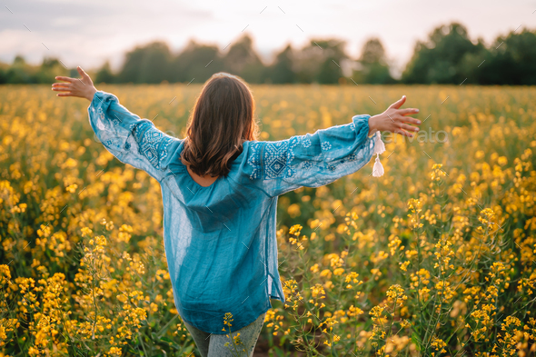 Woman standing with open arms in yellow canola field. Happy free lady in blue embroidered blouse.