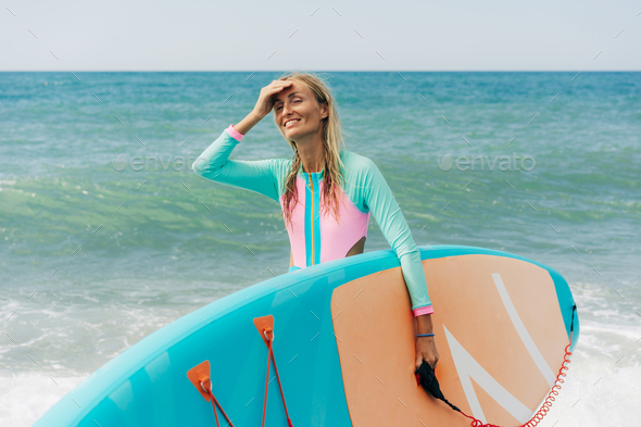 Young happy athletic blonde woman stands on the beach and holds SUP surf.