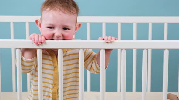 Sad toddler baby boy in the crib gnawing a wooden bars, blue studio background